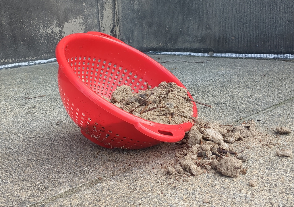 Ein rotes Plastiksieb liegt auf Steinboden und ist halb mit Sand gefüllt